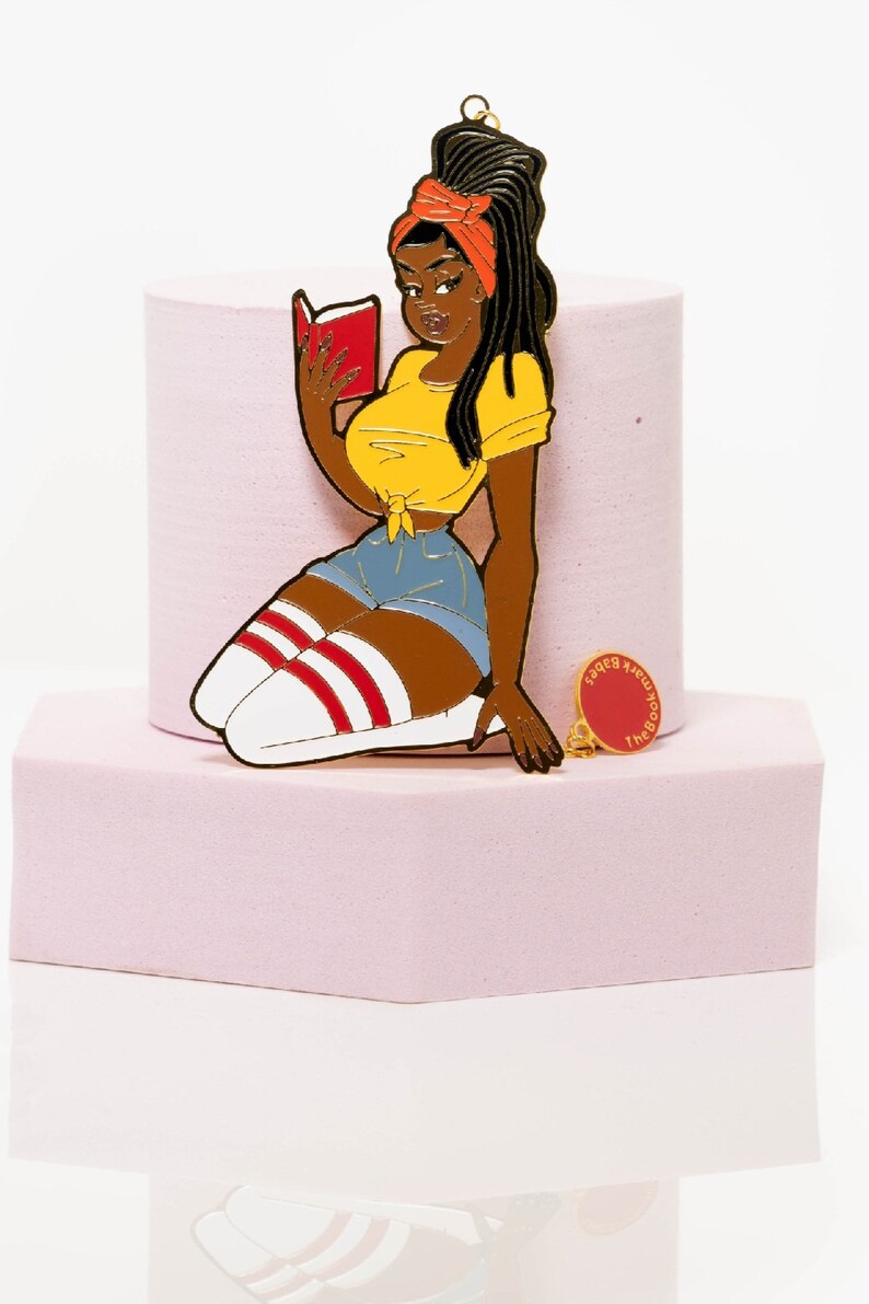 May include: A colorful enamel pin featuring a cartoon woman reading a book, wearing a yellow top, denim shorts, and knee-high socks with red stripes. A smaller pin with the text "The Bookmark Babes" is also present.