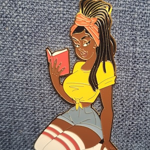 May include: A gold-colored metal pin depicting a cartoon woman with dark skin, wearing a yellow top, denim shorts, and knee-high socks with red stripes. She is reading a red book. The pin is on a blue textured background.