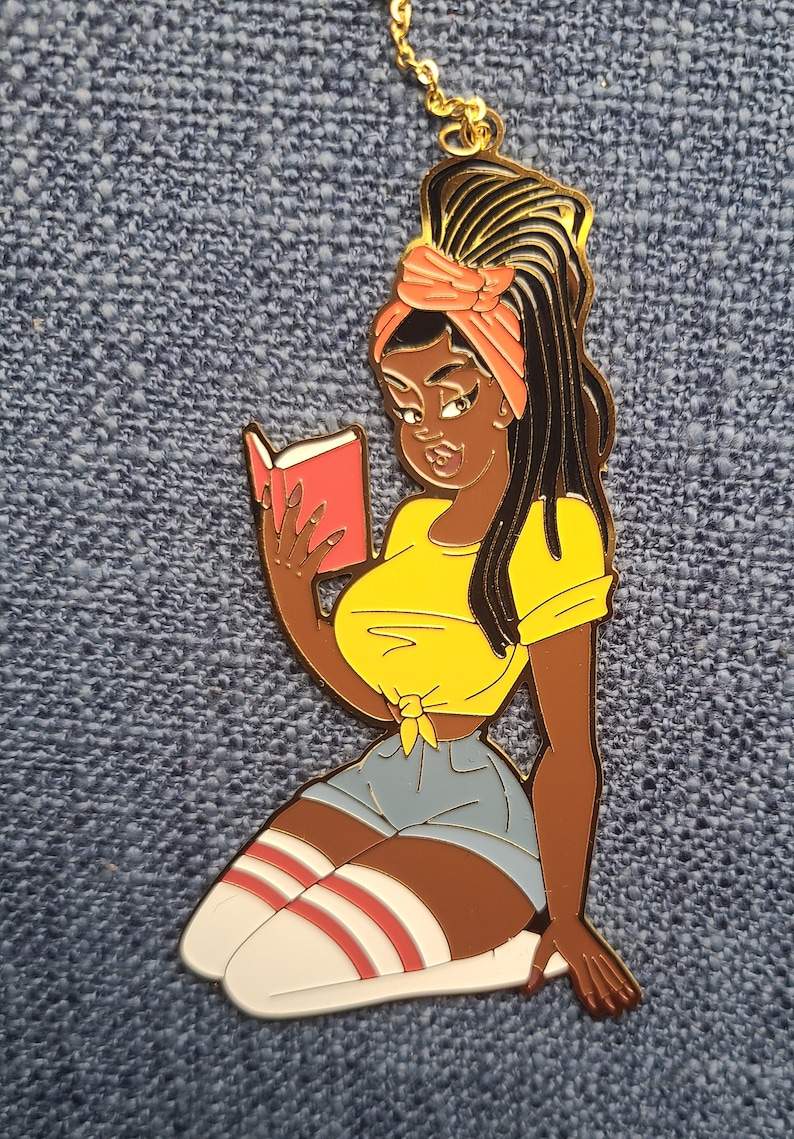 May include: A gold-colored metal pin depicting a cartoon woman with dark skin, wearing a yellow top, denim shorts, and knee-high socks with red stripes. She is reading a red book. The pin is on a blue textured background.