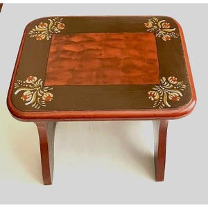 May include: A small, wooden footstool with a rectangular top and curved legs. The top is painted with a reddish-brown center, surrounded by a dark brown border. Floral designs in white, yellow, and orange adorn the corners.