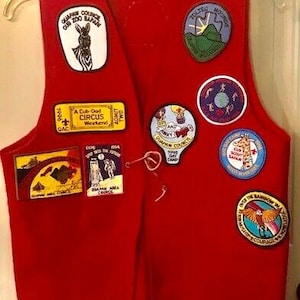 May include: A red vest with multiple patches, including a zebra, a mountain scene, and text that reads "Cub Zoo Safari." The vest is hanging on a clear hanger.