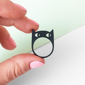 Puede incluir: Anillo negro en forma de gato con grandes ojos blancos. El anillo está hecho de un material negro mate liso.