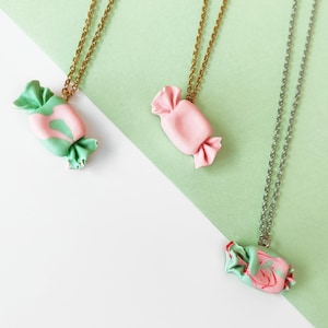 Peut inclure: Trois colliers avec des pendentifs en forme de bonbons. Deux ont des chaînes dorées et un a une chaîne argentée. Les pendentifs sont roses et verts, avec un design enveloppé. Le fond est blanc et vert clair.