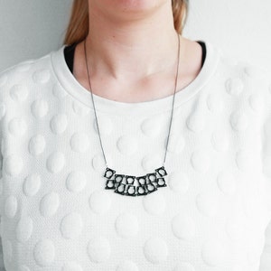 Op de afbeelding: Een zilveren ketting met een geometrisch ontwerp. De ketting is gemaakt van meerdere met elkaar verbonden vierkanten.