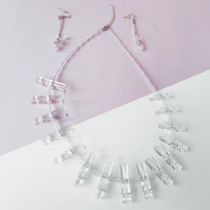 Op de afbeelding: Een heldere, geometrische ketting en bijpassende oorbellen set. De ketting heeft rechthoekige, transparante kralen geregen op een dunne, zilverkleurige draad. De oorbellen hebben een vergelijkbaar ontwerp, met kleine, heldere rechthoekige kralen.