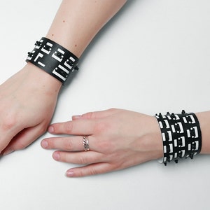 Op de afbeelding: Twee zwarte leren armbanden met een wit geometrisch patroon en zwarte studs. De armbanden worden gedragen op de polsen van een persoon.