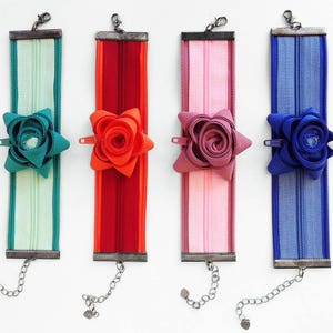 Op de afbeelding: Vier kleurrijke ritsarmbanden. Elke armband heeft een andere kleur: teal, rood, roze en blauw. Elke armband heeft een bijpassend bloemaccent en een zilveren ketting. De armbanden worden op een witte achtergrond gepresenteerd.