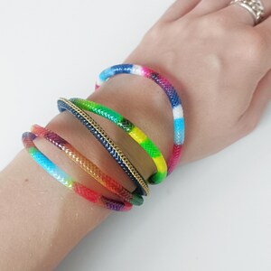 Op de afbeelding: Een verzameling kleurrijke armbanden. Eén armband heeft een regenboog aan kleuren, een andere is groen en geel, en een derde is donkerblauw met een gouden ketting. De armbanden zijn op een pols gestapeld.