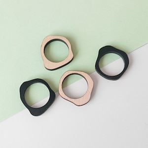 Op de afbeelding: Vier houten ringen, twee zwart en twee naturel hout, met onregelmatige, organische vormen. De ringen zijn gerangschikt op een groen en wit oppervlak.