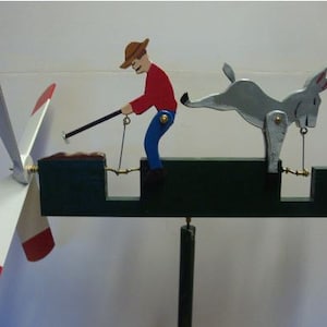 May include: A wooden wind spinner with a red, white, and blue propeller. A man in a brown hat and red shirt is pulling a rope attached to a gray donkey. The donkey is kicking its back leg over a fence.