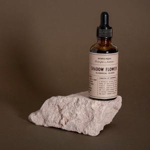 May include: A brown glass bottle with a black dropper top, labeled "Shadow Flower Alchemical Elixir," sits on a light pink rock. The bottle has a cream-colored label with black text. The background is a solid brown.