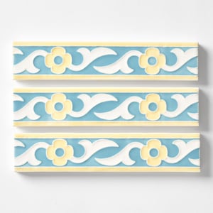 May include: Three decorative ceramic border tiles. Each tile features a repeating pattern of white scrollwork, yellow flowers, and a light blue background. The tiles are rectangular and have a glossy finish.
