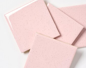 May include: A collection of square pink tiles with a speckled pattern. The tiles are arranged at different angles, showcasing their glossy surface and light pink color. The edges are clean and the overall aesthetic is modern.