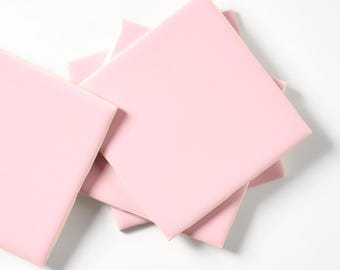 May include: A stack of square, light pink ceramic tiles. The tiles are arranged in a slightly overlapping pattern, with a smooth, matte finish. The edges are clean and the color is consistent across all tiles.