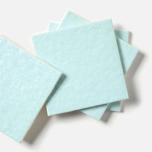 May include: A stack of square ceramic tiles in a light, pale blue color. Each tile features a smooth, glossy finish with a subtle, slightly uneven, and textured surface that catches the light, creating a soft, reflective appearance.