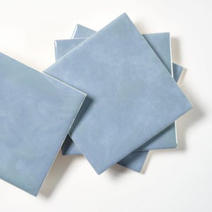 May include: A collection of square, light blue ceramic tiles. The tiles are arranged in a slightly overlapping pattern, showcasing their smooth, glossy surface. The color is a soft, muted blue, suitable for home decor projects.