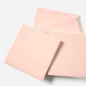 May include: Four square, pale pink ceramic tiles are arranged on a white surface. The tiles have a glossy finish and are slightly overlapping, showcasing their smooth, clean edges. The tiles are a soft, pastel color.
