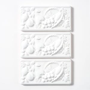 May include: Three white rectangular ceramic tiles with a raised relief design. Each tile features a basket overflowing with grapes, pears, and lemons, surrounded by floral accents. The tiles are arranged vertically against a white background.