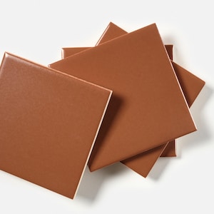 May include: A collection of square, terracotta-colored tiles with a glossy finish. The tiles are arranged in a slightly overlapping pattern, showcasing their smooth surfaces and clean edges. The tiles are approximately 12 inches by 12 inches.