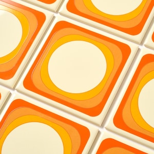 May include: Square ceramic tiles with a retro design. Each tile features a central off-white circle surrounded by concentric squares in shades of orange and yellow. The tiles are arranged in a grid pattern, creating a repeating geometric design.
