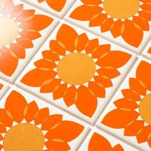 May include: Square white tiles with a repeating pattern of orange and yellow sunflowers. The sunflower design features a central yellow circle surrounded by orange petals. The tiles are arranged in a grid.
