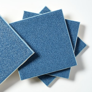 May include: A collection of square, blue ceramic tiles with a textured surface. The tiles are arranged in a slightly overlapping pattern, showcasing their color and texture. The tiles have a glossy finish and are suitable for various home improvement projects.