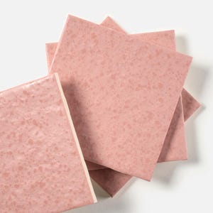 May include: A stack of square, pink ceramic tiles with a speckled pattern. The tiles are arranged in a slightly overlapping manner, showcasing their edges and surface texture. The tiles have a glossy finish.