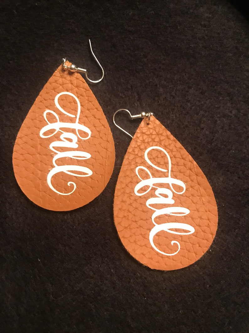 May include: A pair of brown leather teardrop earrings with the word "fall" written in white script on each earring.