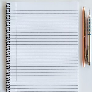 Op de afbeelding: Een wit, gestreept notitieboek met een zwarte spiraalbinding. Twee pennen, een gouden en een zilveren, liggen naast het notitieboek.