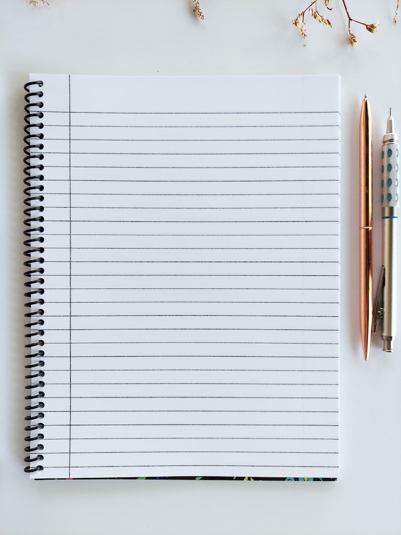 Op de afbeelding: Een wit, gestreept notitieboek met een zwarte spiraalbinding. Twee pennen, een gouden en een zilveren, liggen naast het notitieboek.