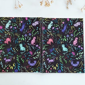 Op de afbeelding: Een zwart spiraalnotitieblok met een kleurrijk patroon van katten en bloemen. De katten zijn in verschillende kleuren, waaronder roze, paars, blauw en groen. De bloemen zijn in tinten geel, roze en groen.