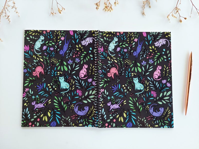 Op de afbeelding: Een zwart spiraalnotitieblok met een kleurrijk patroon van katten en bloemen. De katten zijn in verschillende kleuren, waaronder roze, paars, blauw en groen. De bloemen zijn in tinten geel, roze en groen.