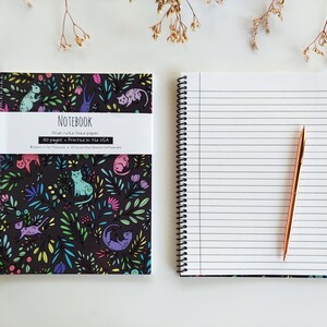 Op de afbeelding: Een zwart notitieboek met een kleurrijk katten- en bloemenpatroon op de omslag. Het notitieboek heeft 80 pagina's met breed geruit papier en is gedrukt in de VS. Het notitieboek is open om een lege pagina te laten zien met een gouden pen erop.