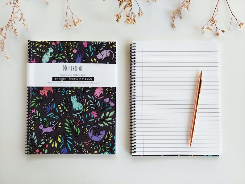 Op de afbeelding: Een zwart notitieboek met een kleurrijk katten- en bloemenpatroon op de omslag. Het notitieboek heeft 80 pagina's met breed geruit papier en is gedrukt in de VS. Het notitieboek is open om een lege pagina te laten zien met een gouden pen erop.