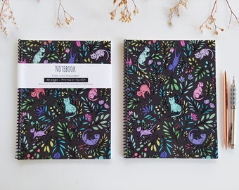 Op de afbeelding: Twee zwarte spiraalnotitieblokken met kleurrijke katten- en bloemendesigns. De notitieblokken hebben de tekst "NOTEBOOK Breed geruit papier 50 pagina's - Gedrukt in de VS" op de voorzijde.
