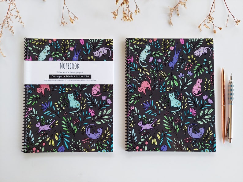 Op de afbeelding: Twee zwarte spiraalnotitieblokken met kleurrijke katten- en bloemendesigns. De notitieblokken hebben de tekst "NOTEBOOK Breed geruit papier 50 pagina's - Gedrukt in de VS" op de voorzijde.