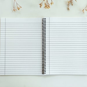 Op de afbeelding: Een open spiraalnotitieblok met witte, gestreepte pagina's en een zwarte plastic binding. Een roosgoudkleurige pen ligt op een wit oppervlak naast het notitieblok.