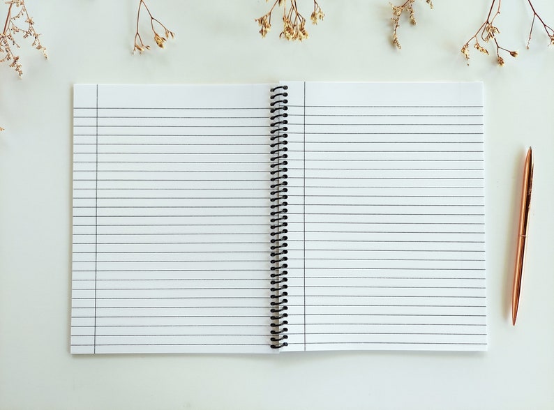 Op de afbeelding: Een open spiraalnotitieblok met witte, gestreepte pagina's en een zwarte plastic binding. Een roosgoudkleurige pen ligt op een wit oppervlak naast het notitieblok.
