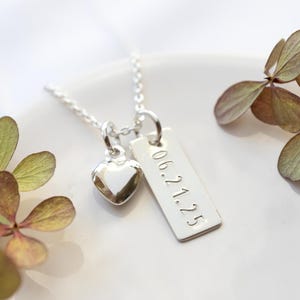 May include: A silver necklace with a heart-shaped charm and a rectangular tag. The tag is engraved with the date "06.21.25". The necklace is displayed on a white surface with dried flowers in the background.