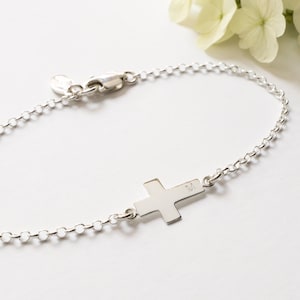 May include: A silver chain bracelet with a small silver cross charm. The cross has the letter "M" engraved on it.
