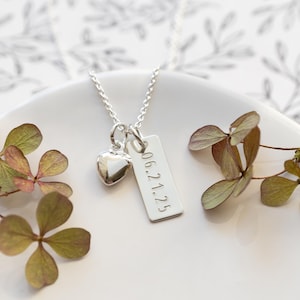 K&ouml;nnte beinhalten: Eine silberne Halskette mit einem Herzanh&auml;nger und einem rechteckigen Anh&auml;nger mit der Gravur "06.21.25". Die Kette liegt auf einer wei&szlig;en Oberfl&auml;che, mit getrockneten Blumen.