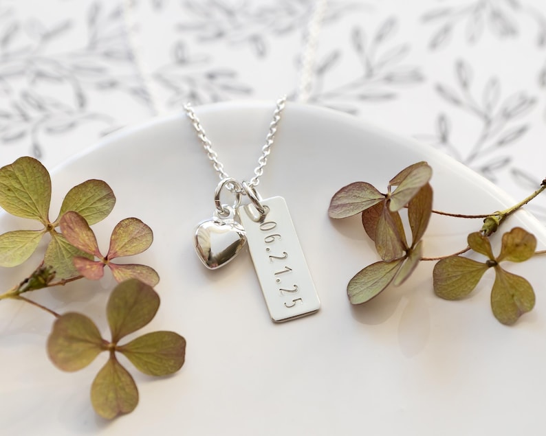 K&ouml;nnte beinhalten: Eine silberne Halskette mit einem Herzanh&auml;nger und einem rechteckigen Anh&auml;nger mit der Gravur "06.21.25". Die Kette liegt auf einer wei&szlig;en Oberfl&auml;che, mit getrockneten Blumen.