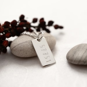 May include: Silver necklace with a rectangular tag engraved with the numbers 57751. The necklace is displayed on a white surface with two gray stones.