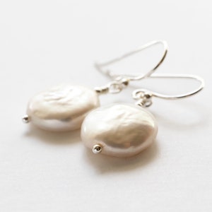 May include: A pair of elegant earrings featuring large, off-white, coin-shaped pearls. The pearls are suspended from delicate silver-colored ear wires. The earrings have a simple, classic design, perfect for everyday wear or special occasions.