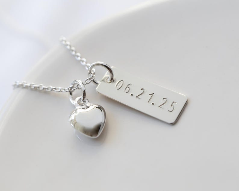 K&ouml;nnte beinhalten: Eine silberne Halskette mit einem Herzanh&auml;nger und einem rechteckigen Anh&auml;nger. Auf dem Anh&auml;nger ist das Datum "06.21.25" eingraviert. Die Halskette liegt auf einer wei&szlig;en Oberfl&auml;che.