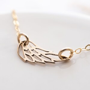 May include: A gold-toned necklace featuring a delicate, openwork wing pendant suspended from a fine chain. The wing design includes cut-out details that create a stylized feather pattern, with the chain attached to two small jump rings on the pendant.