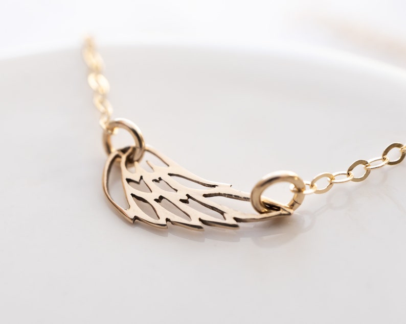 May include: A gold-toned necklace featuring a delicate, openwork wing pendant suspended from a fine chain. The wing design includes cut-out details that create a stylized feather pattern, with the chain attached to two small jump rings on the pendant.