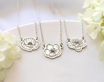 May include: Three silver-toned necklaces featuring flat, irregular-shaped metal pendants. Each pendant is engraved with a distinct black line-art floral design, including a poppy, a cherry blossom, and a water lily, attached to a delicate silver chain.