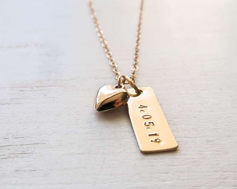 K&ouml;nnte beinhalten: Eine goldfarbene Halskette mit einem kleinen Herzanh&auml;nger und einem rechteckigen Anh&auml;nger. Der Anh&auml;nger ist mit dem Datum "4.05.19" graviert. Kette und Anh&auml;nger haben eine gl&auml;nzende Oberfl&auml;che. Der Hintergrund ist eine helle Holzoberfl&auml;che.