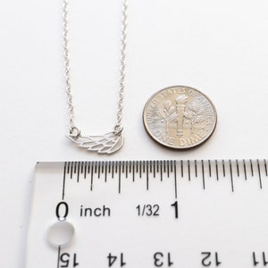 May include: A silver-toned necklace featuring a small wing-shaped pendant. The pendant is positioned next to a United States dime and a clear plastic ruler, showing the wing charm measures approximately 0.75 inches in total width.